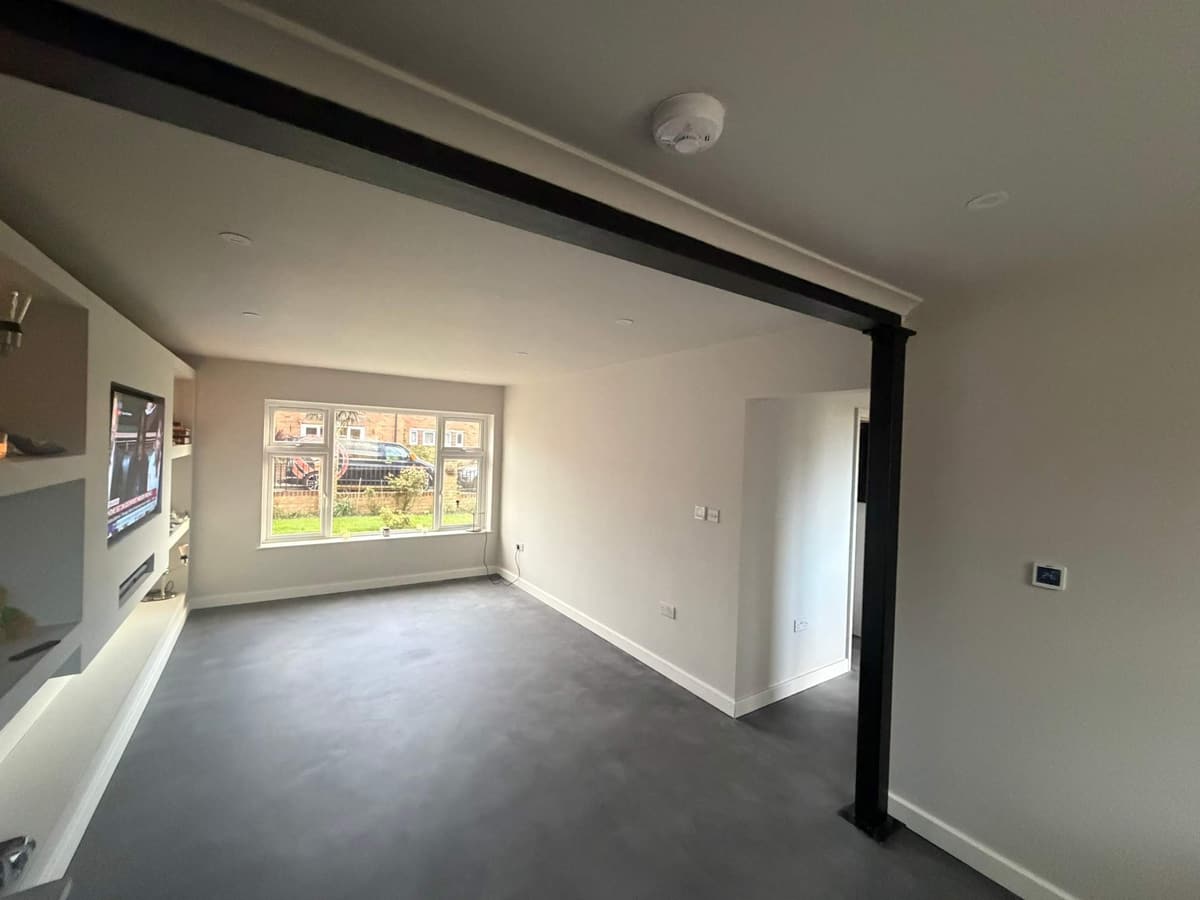 After: the same lounge finished, with smooth white walls, a black steel beam, a painted media wall and a grey floor
